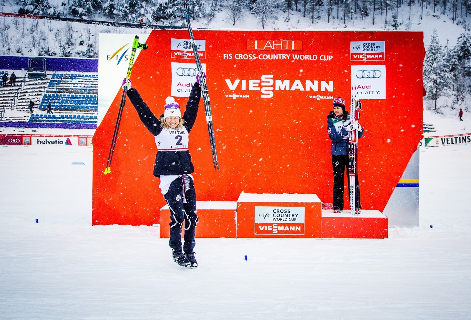 Two people stand on podiums during a snowy FIS Cross-Country World Cup event. The individual on the taller podium, labeled "1," raises their arms and poles in triumph. The other figure stands on a lower podium, labeled "2." A vibrant orange backdrop proudly displays sponsor names, including Vitodens boilers by Viessmann.