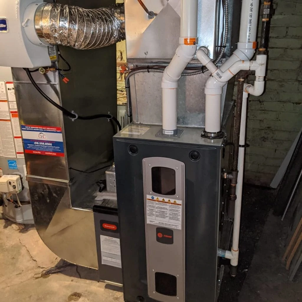 A Trane HVAC unit installed in a basement ensures optimal heating and cooling. The unit features connected ducts, white pipes, and a metal rectangular vent above. Cement walls and other fixtures complete the City Home Comfort setup.