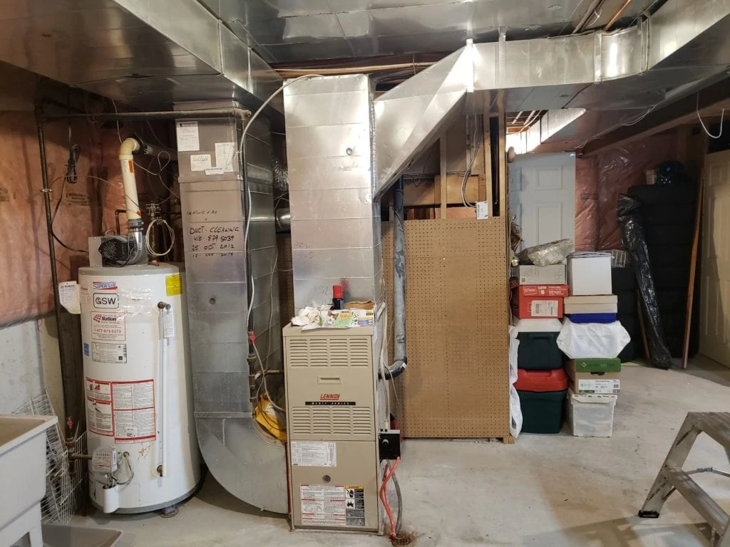 The basement area features a furnace and water heater connected by ducts, surrounded by pipes. Shelving with assorted bins line the right side, while various construction materials lie scattered. Insulation and a pink vapor barrier adorn the walls, preparing the space for Fall. The ceiling remains unfinished.