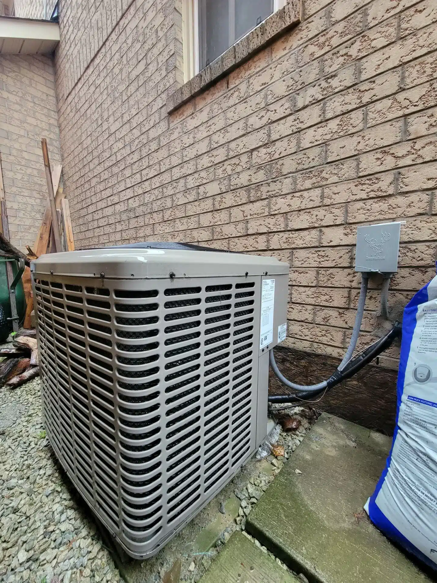 Outdoor air conditioning unit on a concrete slab next to a brick wall. Electrical wiring runs from the unit to a mounted box. A bag of salt or ice melt is leaning against the wall nearby. Gravel and wooden debris are scattered around.
