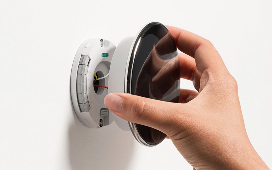 A hand is seen installing a round, 4th Gen Nest Learning Thermostat onto a wall-mounted base. The base has exposed wiring and some internal components visible. The outer front of the thermostat is reflective and sleek, showing a primarily minimalist design, perfect for City Home Comfort.