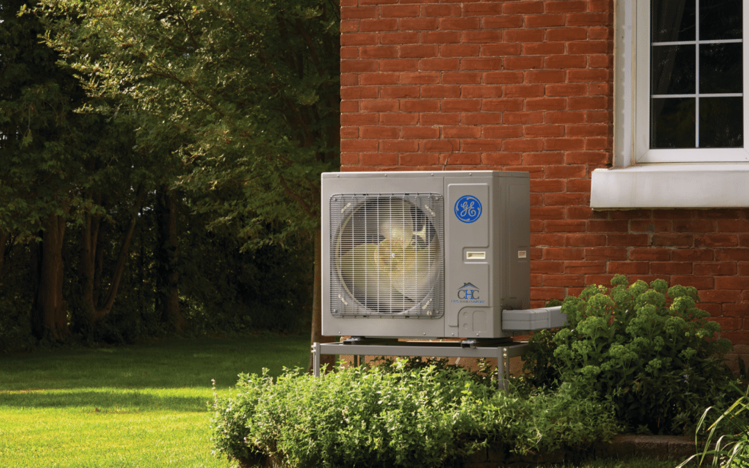 A GE Connect Variable Speed Heat Pump, boasting High-Efficiency Inverter Technology and a capacity between 24,000-60,000 BTU, is positioned beside a brick house surrounded by lush greenery. The unit displays a visible fan and company branding as it rests on a metal platform. A backdrop of trees and grassy lawn completes the scene.