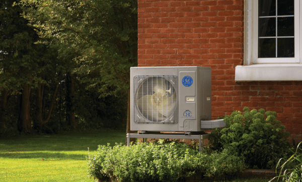 A GE Connect Variable Speed Heat Pump, boasting High-Efficiency Inverter Technology and a capacity between 24,000-60,000 BTU, is positioned beside a brick house surrounded by lush greenery. The unit displays a visible fan and company branding as it rests on a metal platform. A backdrop of trees and grassy lawn completes the scene.