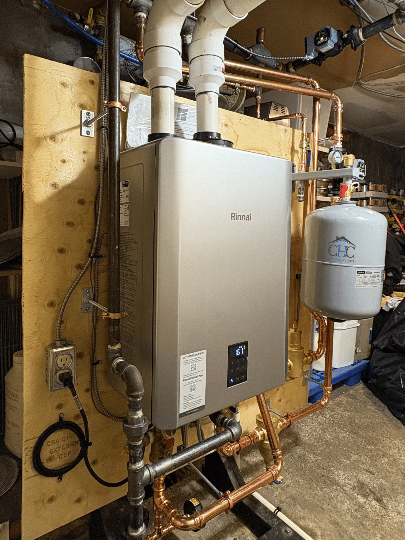 A New Rinnai I-Series Plus Combi and Heat Only Boiler is elegantly installed on a plywood board with copper piping. An expansion tank labeled "CHC" is positioned on the right, encircled by electrical outlets and various plumbing components in a snug basement environment.