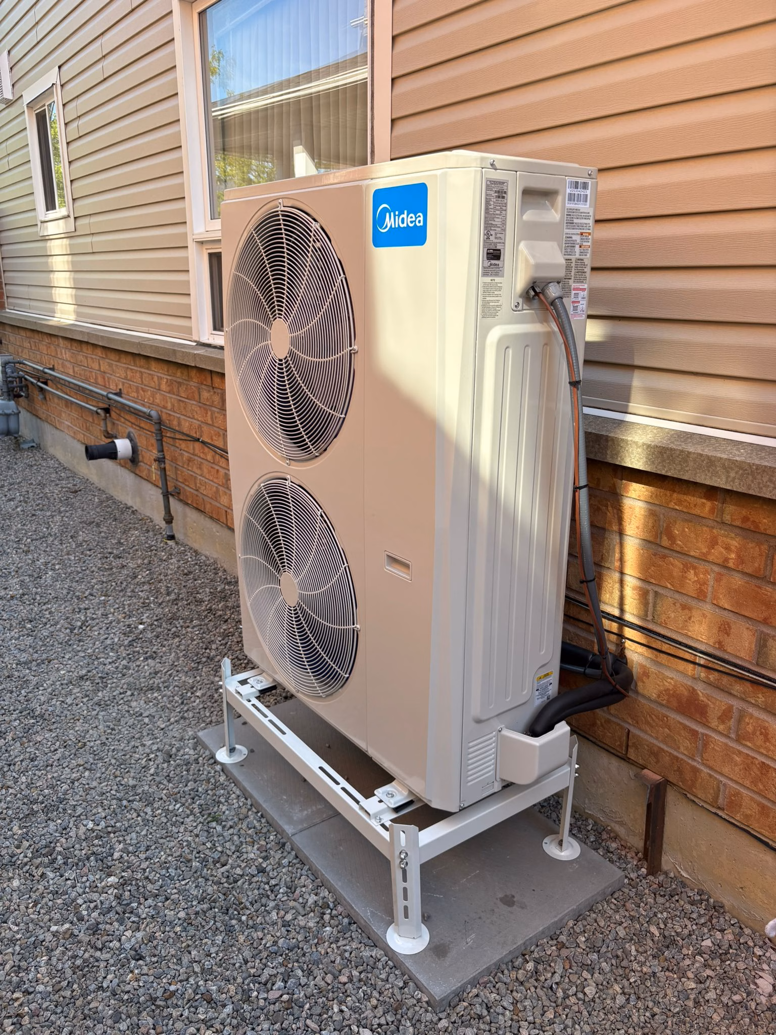 A white Midea outdoor air conditioning unit with two large fans is installed on a metal stand next to a brick and siding house exterior, with gravel covering the ground.