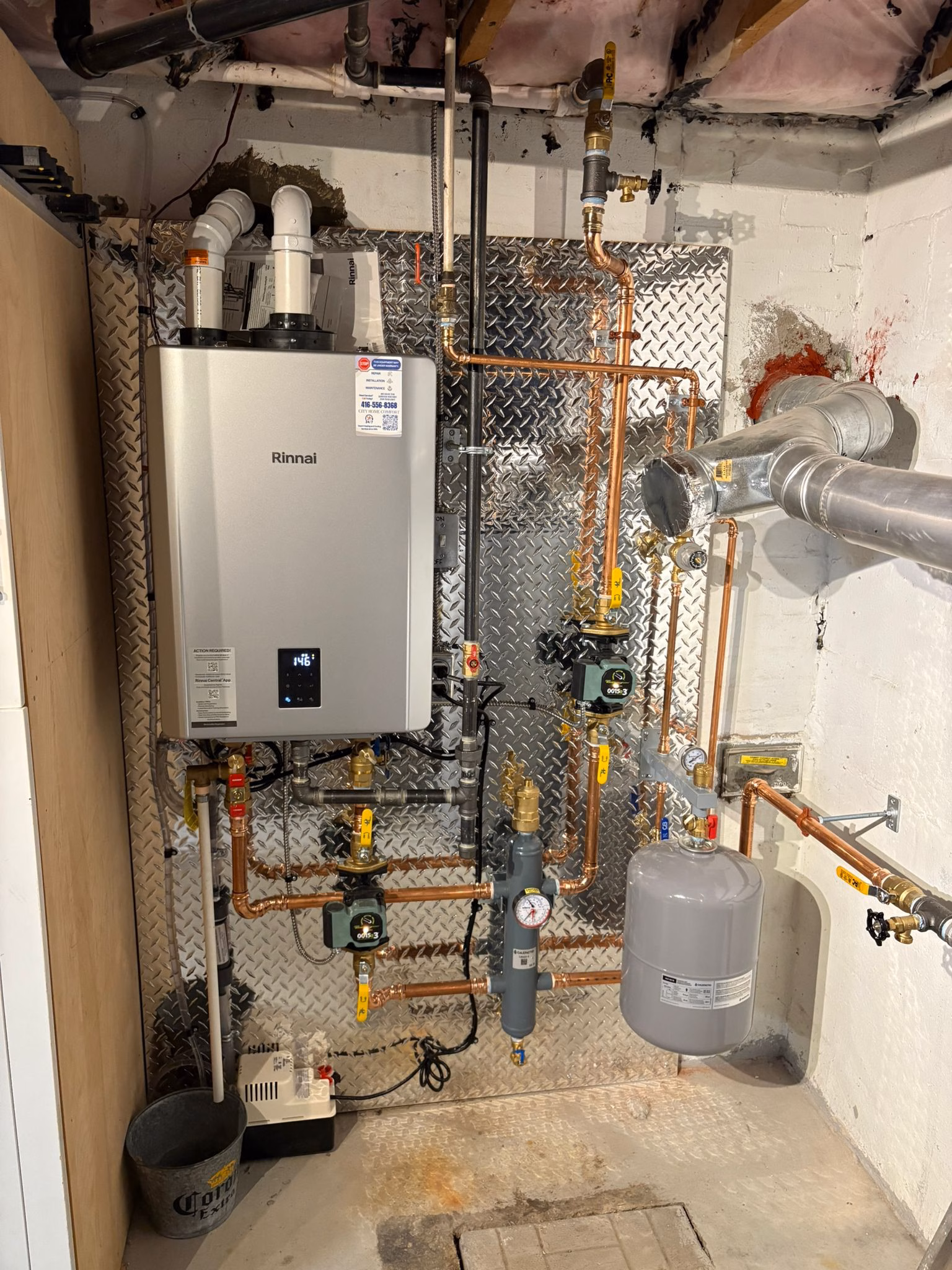 A modern wall-mounted boiler system with copper piping, pressure gauges, valves, and a grey expansion tank in a basement utility area. The concrete walls and exposed ceiling pipes are also visible.