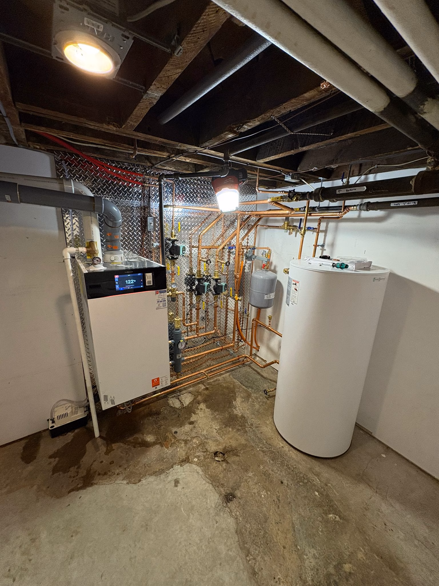 A basement utility room with exposed ceiling beams, a Viessmann Boiler system unit with a digital display, copper pipes, a white cylindrical tank, and a concrete floor with a water stain. A basement utility room with exposed ceiling beams, a Viessmann Boiler system unit with a digital display, copper pipes, a white cylindrical tank, and a concrete floor with a water stain.